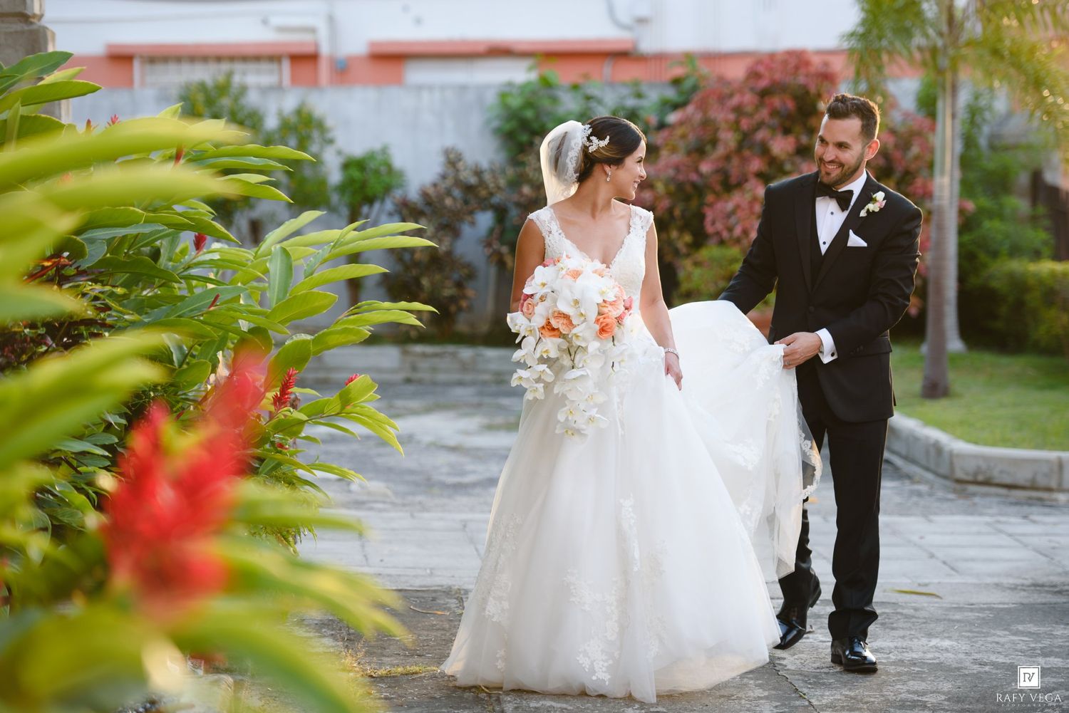 Bride in white ball gown and groom in black tuxedo walking together on a tropical garden path with lush greenery.