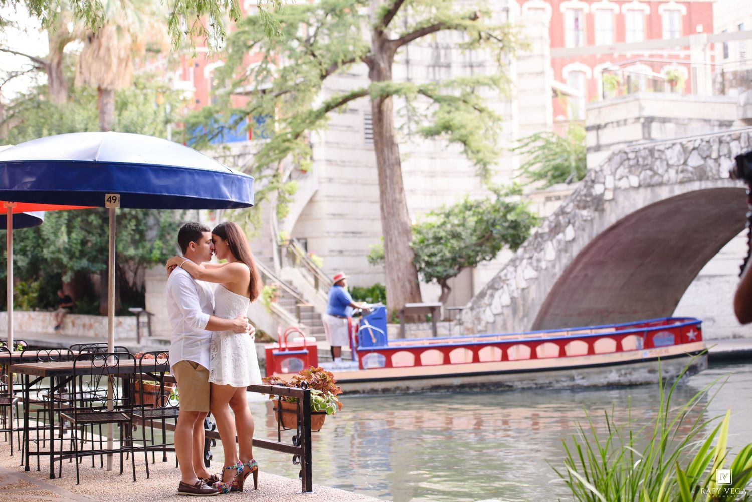 Couple embracing romantically along the San Antonio River Walk with a bridge and tour boat in the background.
