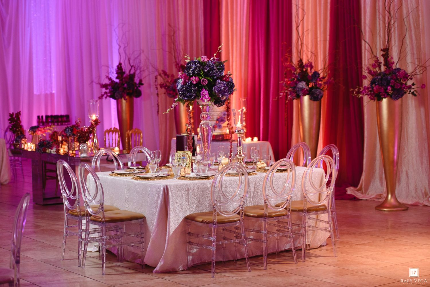 Luxurious wedding reception with gold vases, purple floral centerpieces, and pink draping under romantic lighting.