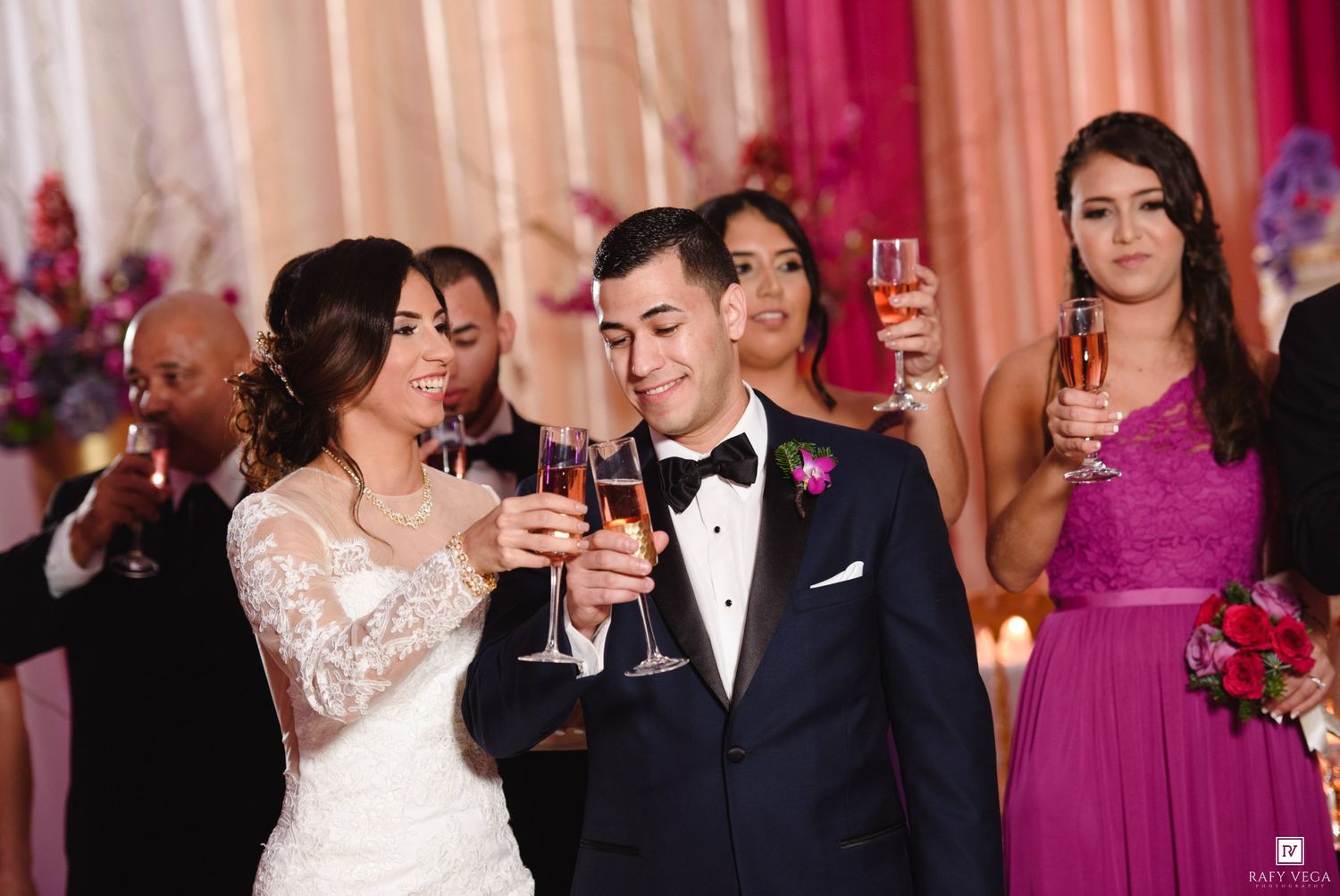 Bride and groom toasting with champagne at wedding reception, surrounded by guests in elegant pink and purple attire.