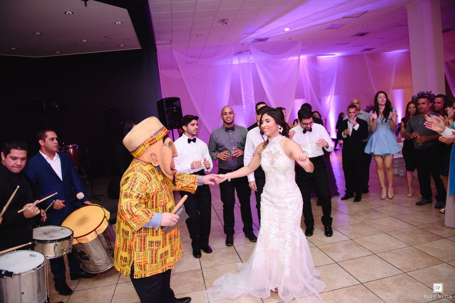 Bride dancing with a musician in a colorful outfit at a wedding reception with guests watching.