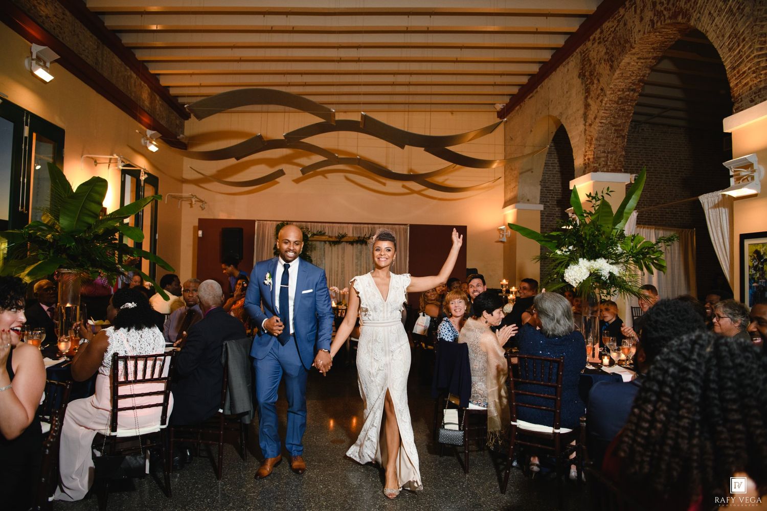Bride and groom make grand entrance at elegant wedding reception venue with guests celebrating around decorated tables.