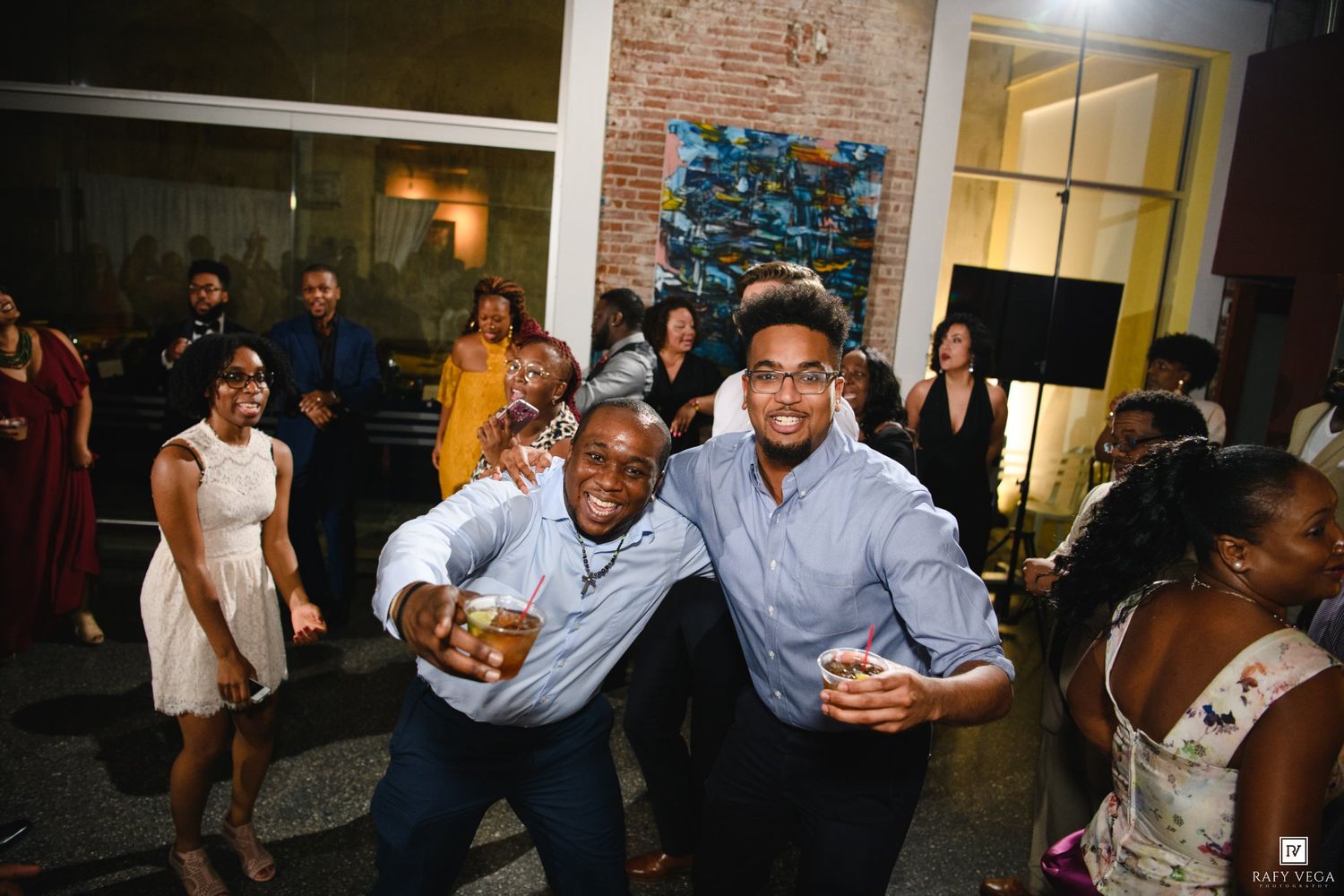 Two men smile and dance together at a lively indoor party with guests celebrating in the background.