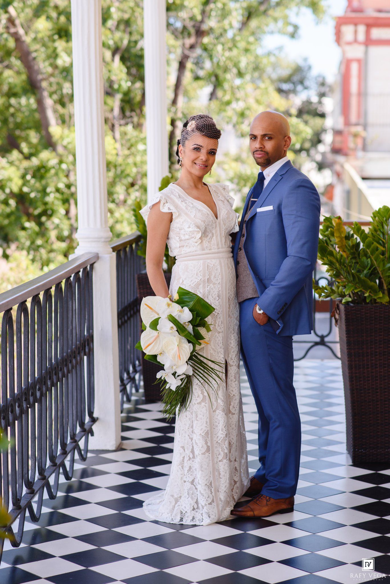 Bride and groom posing on a checkered floor balcony with white columns, bride holding white floral bouquet.