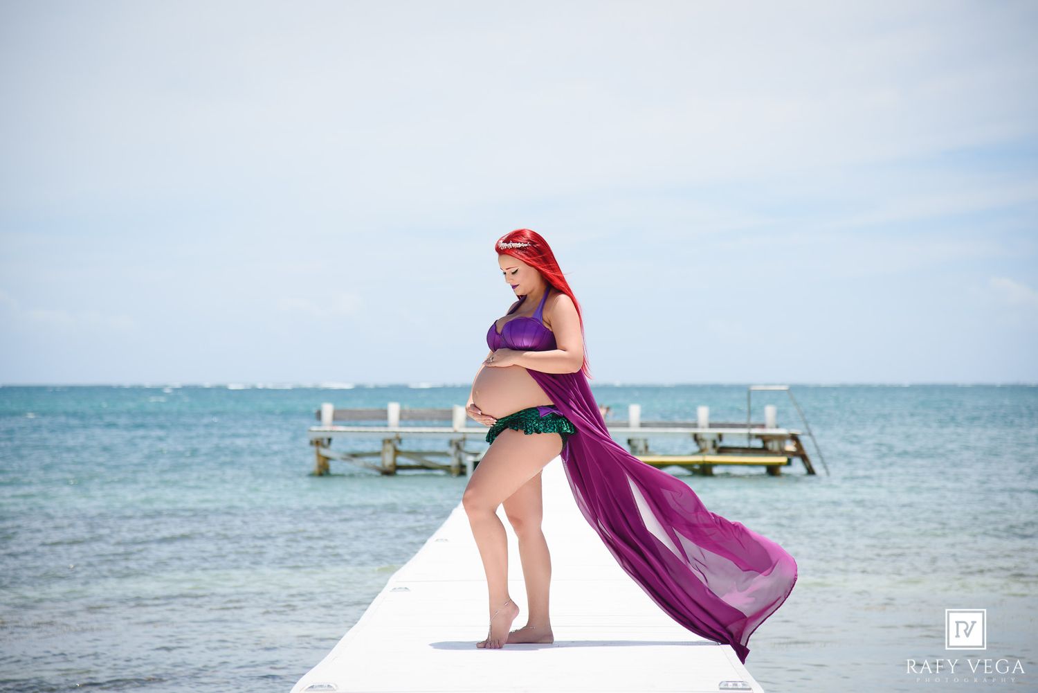 Pregnant woman in mermaid costume with red hair poses on white pier over turquoise ocean waters.