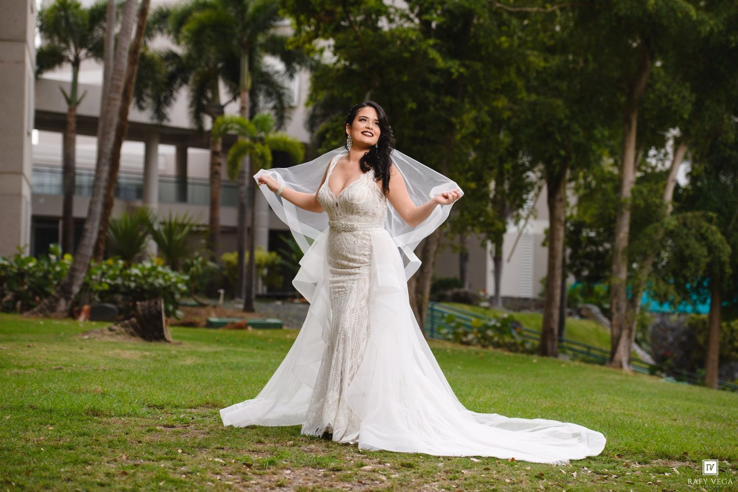 Bride in white lace gown with veil spread wide, posing on green lawn outdoors.