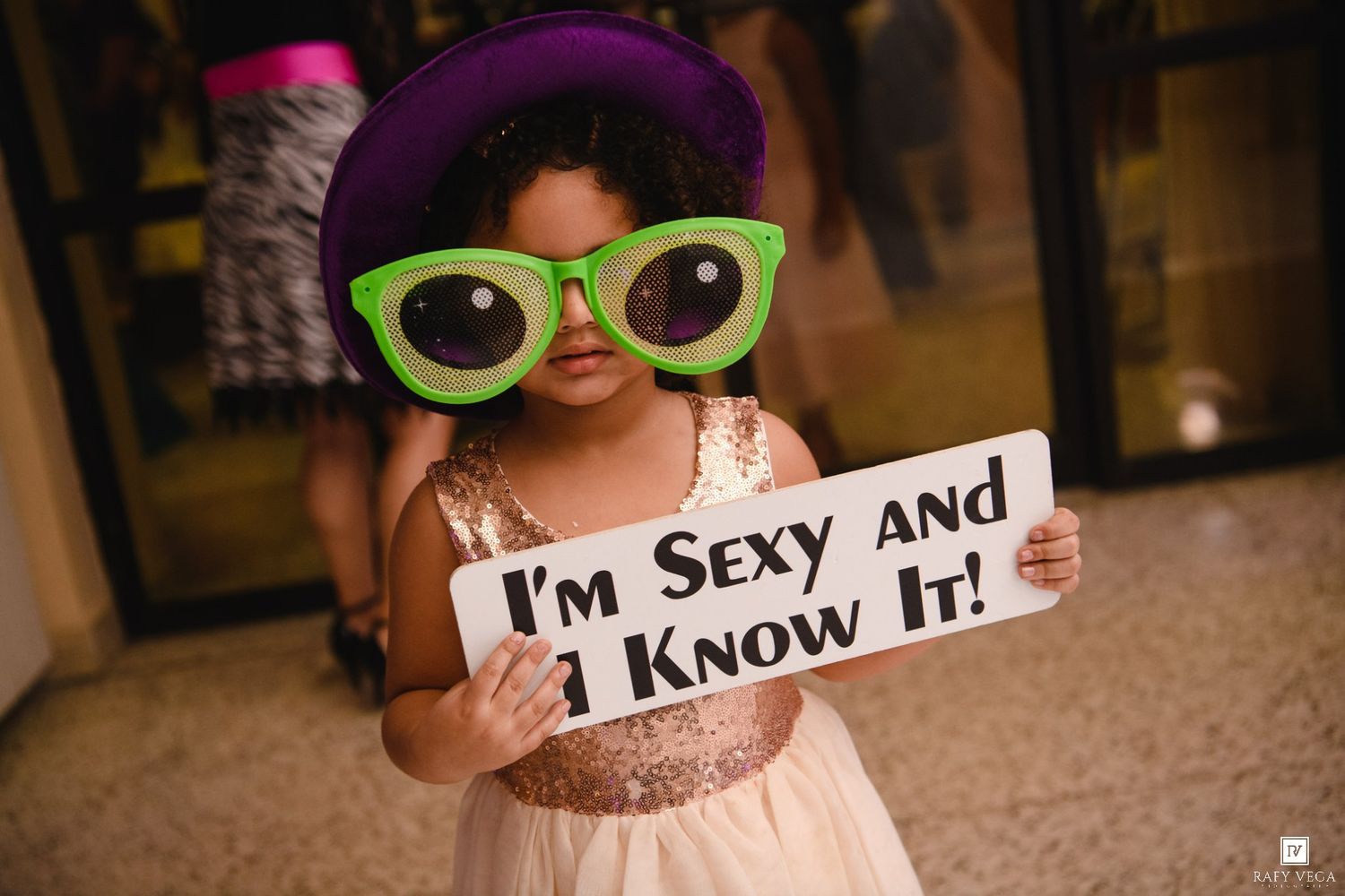 Cute toddler girl wearing oversized green glasses and purple hat holding I'm Sexy and I Know It sign at party.