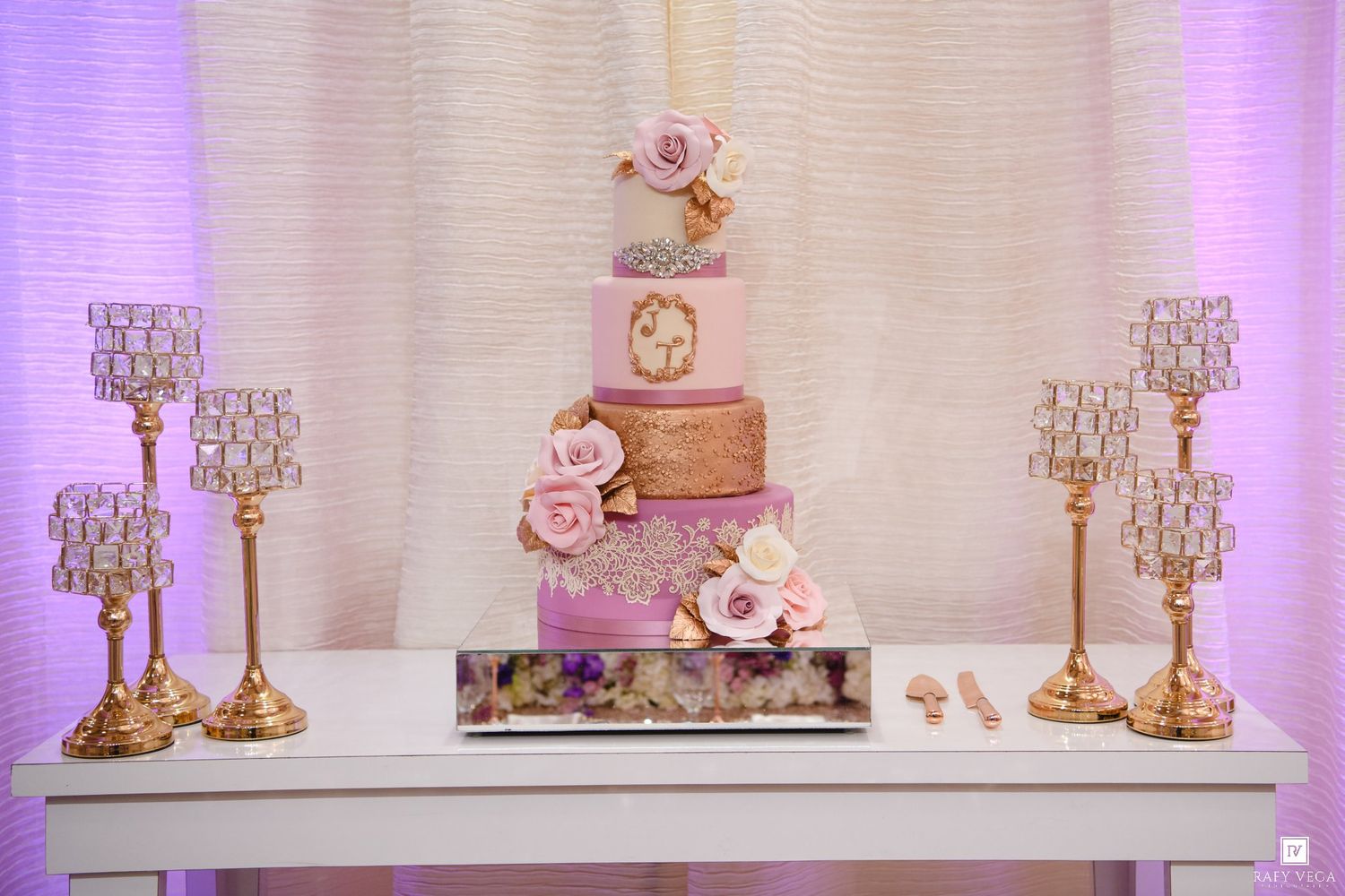 Elegant four-tier pink and gold wedding cake adorned with roses on a white table with gold candelabras.