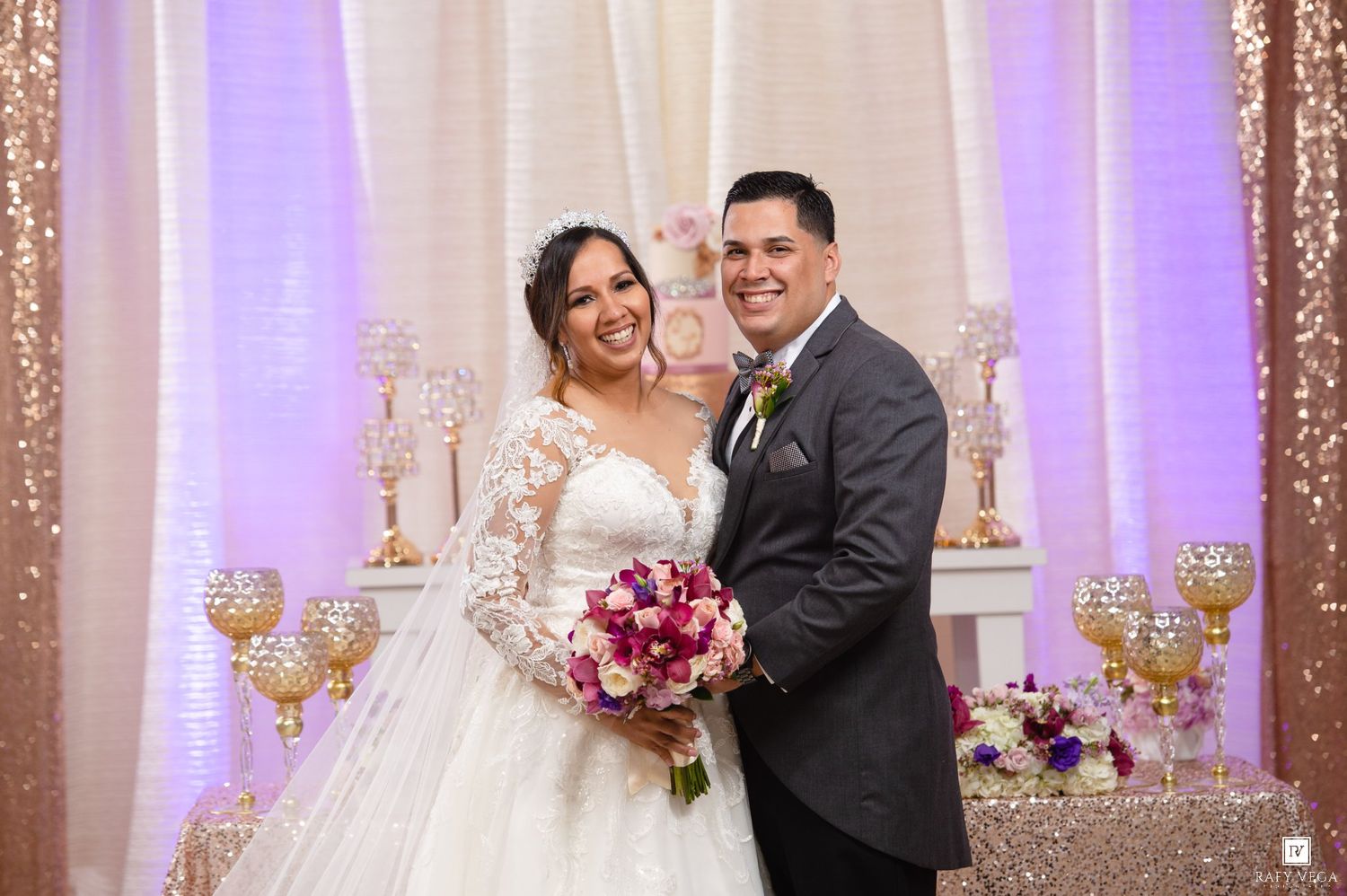 Happy newlyweds pose together smiling at decorated wedding venue with purple flowers and gold accents.