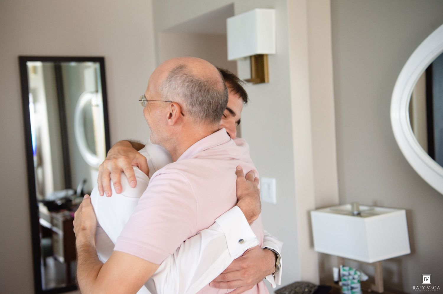 Two men share a warm embrace in a bright room, one wearing glasses and a pink shirt.