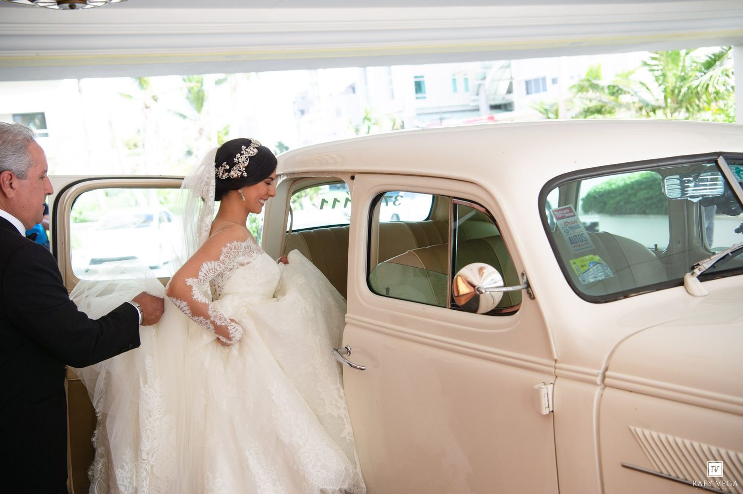 Bride in white gown entering a vintage cream classic car, assisted by a man, at an elegant wedding venue.