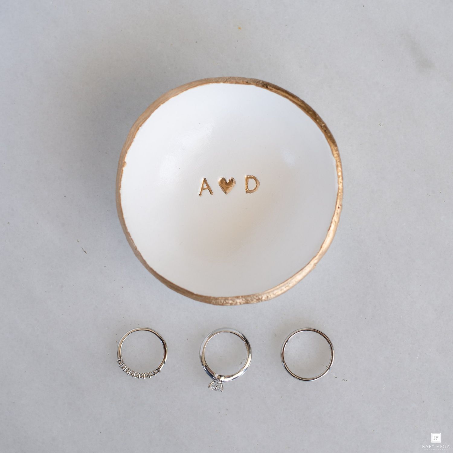 White ceramic ring dish with gold rim and A heart D monogram, displayed with three silver rings on marble surface.