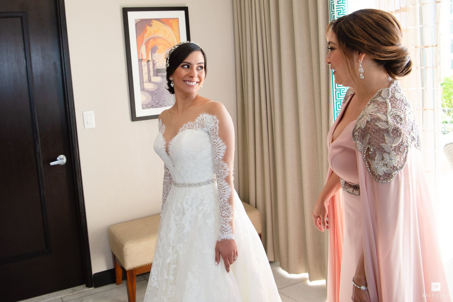 Bride in elegant lace wedding gown admired by woman in pink dress during bridal getting ready moment.