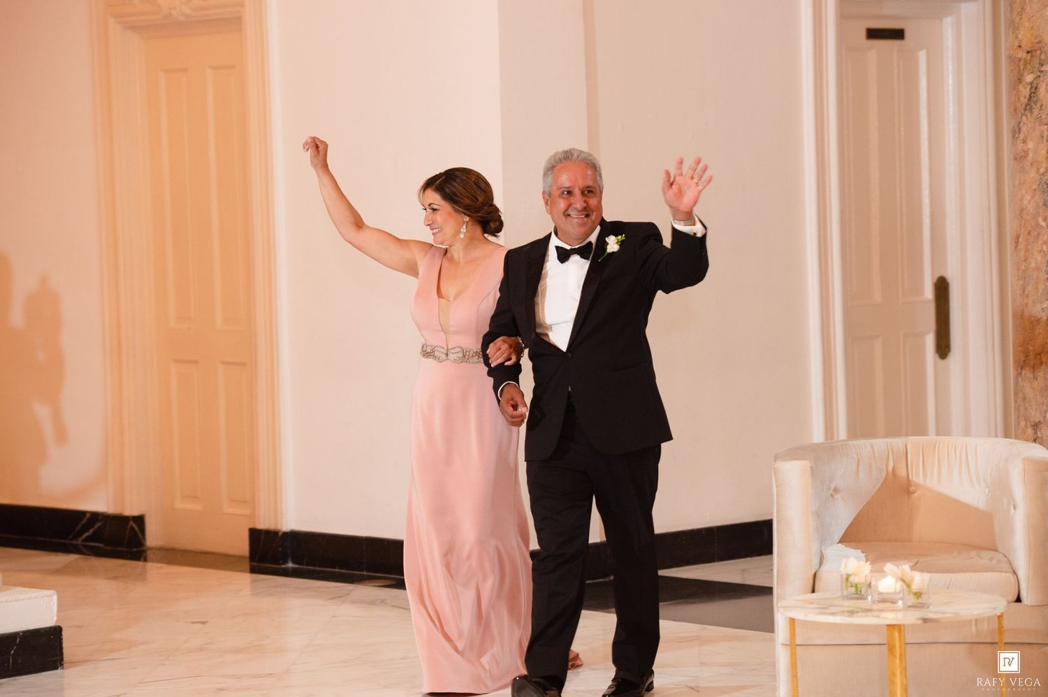 Elegant couple in formal attire waving as they enter a grand ballroom at a wedding reception.