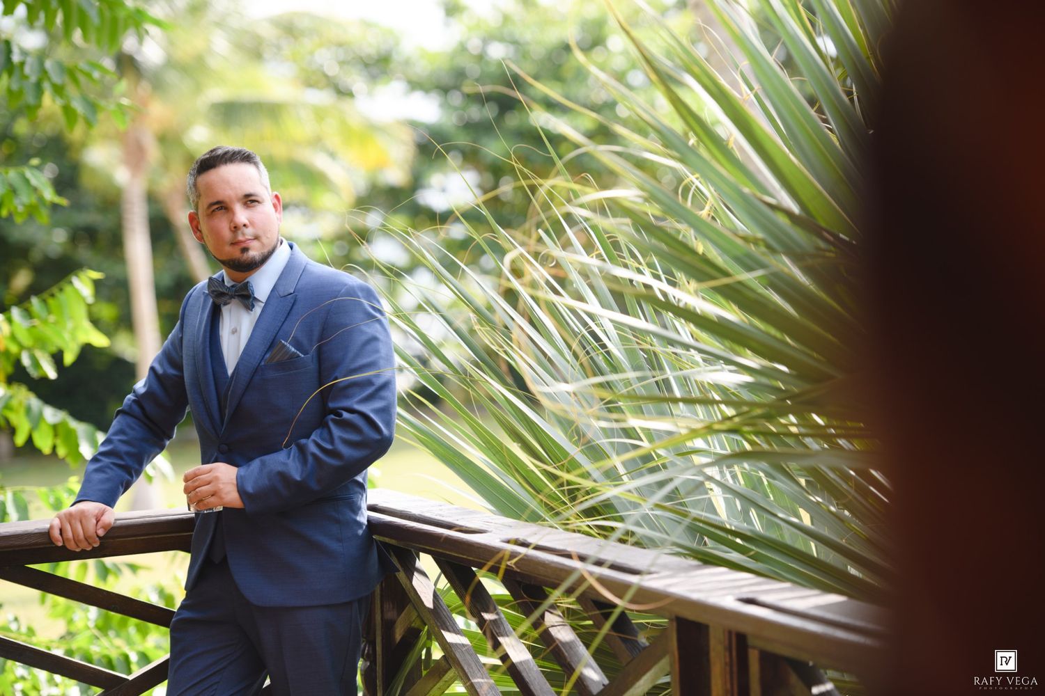 Elegant groom in blue suit with bow tie posing on wooden deck surrounded by tropical palm trees.