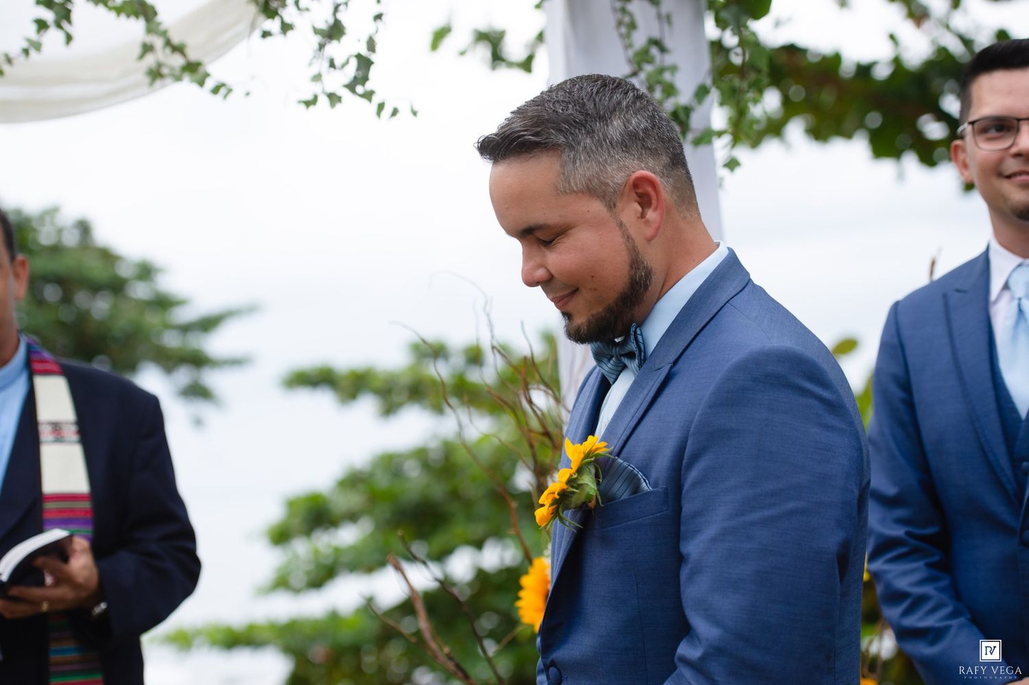 Groom in blue suit with bow tie and sunflower boutonniere smiling during outdoor wedding ceremony.