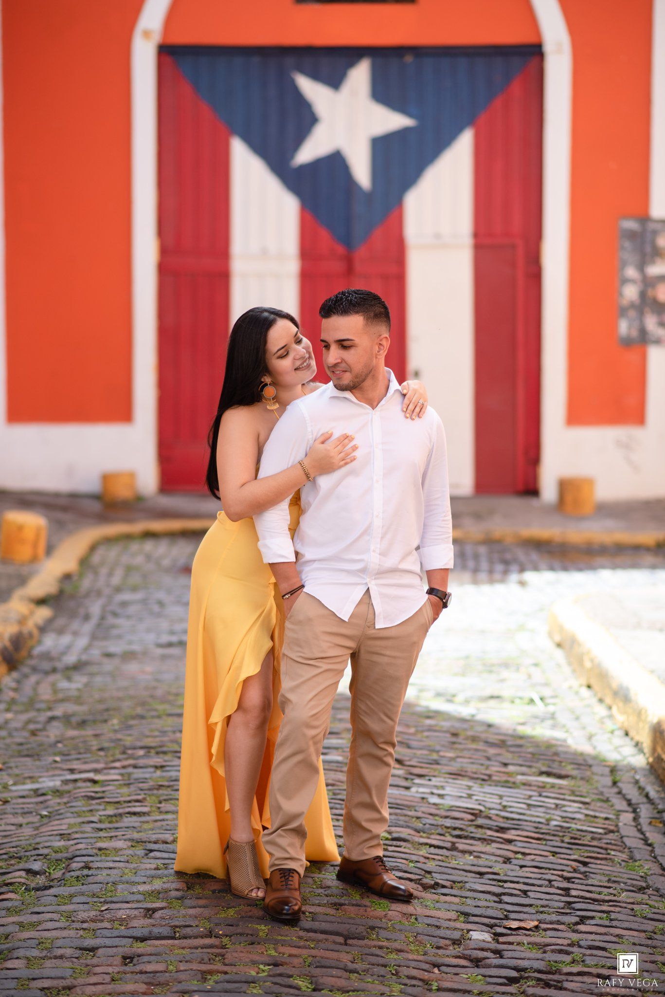 Couple embracing on cobblestone street in Old San Juan, Puerto Rico, with colorful mural of Puerto Rican flag on wall behind them.