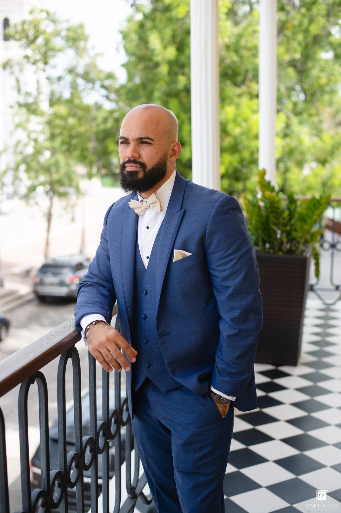 Elegant bald groom with beard poses in navy blue three-piece suit on balcony with checkered floor.