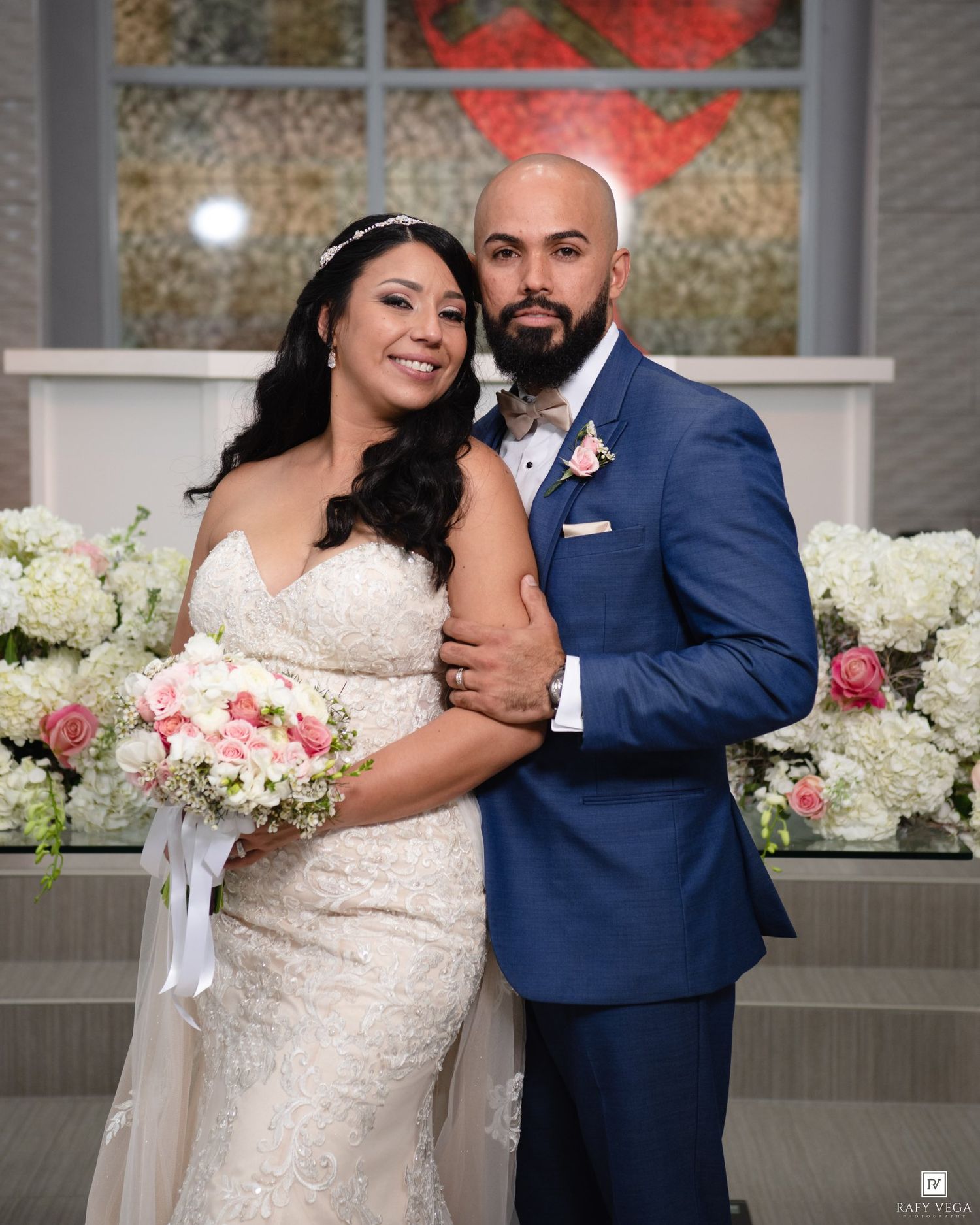 Bride in lace gown holding pink bouquet poses with groom in blue suit at elegant church wedding ceremony.