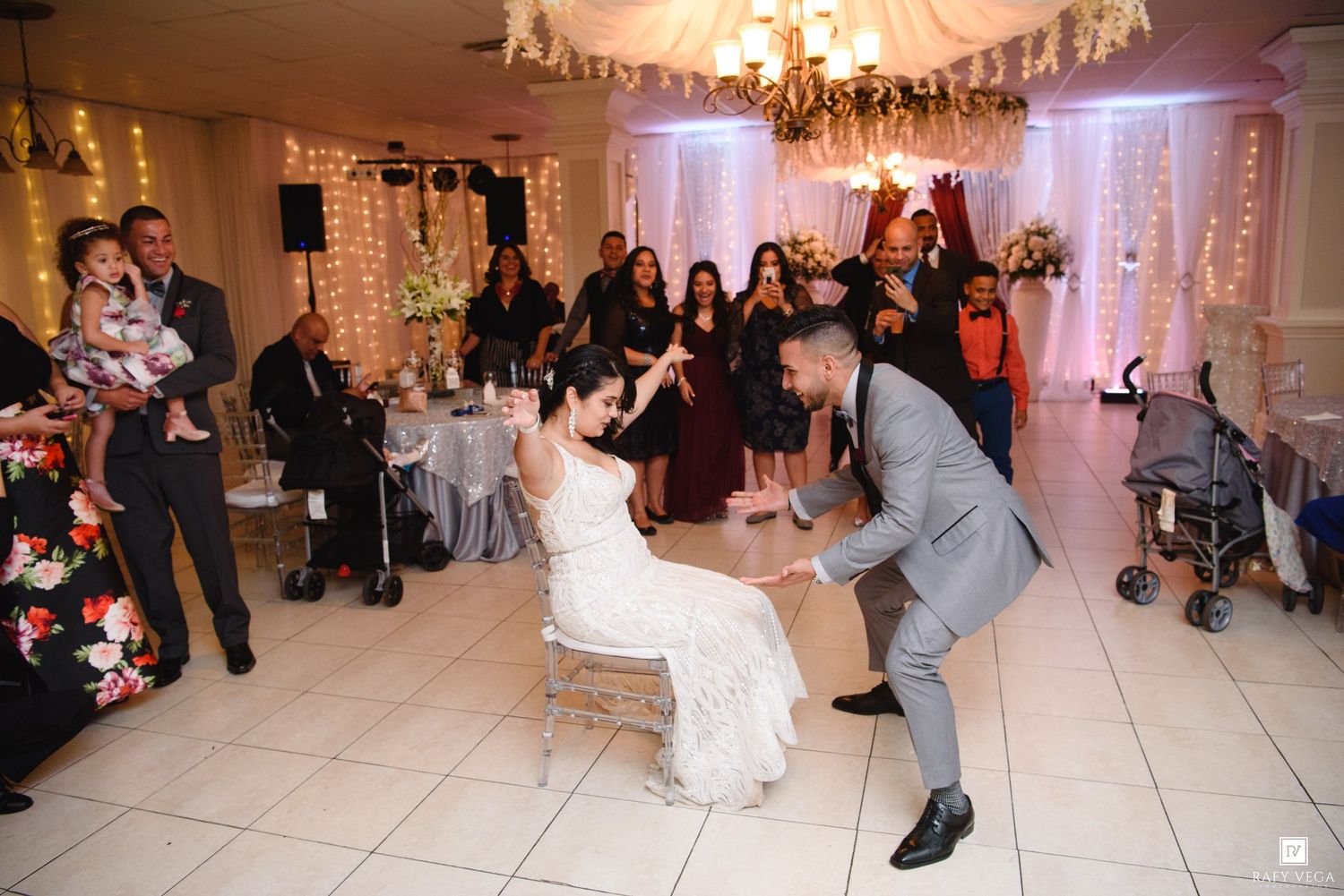 Groom removes garter from bride seated on chair at elegant wedding reception with guests watching.
