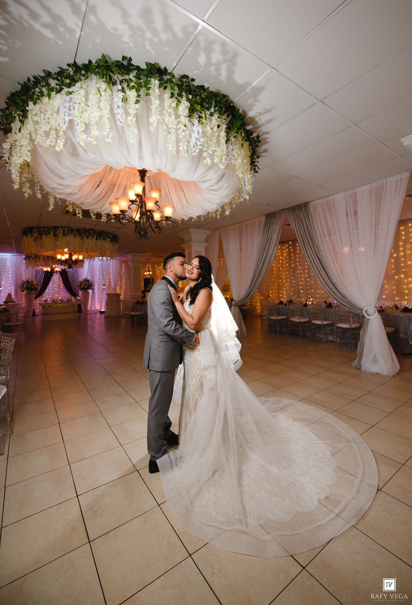 Bride and groom share first dance kiss under floral chandelier in elegant wedding reception venue.