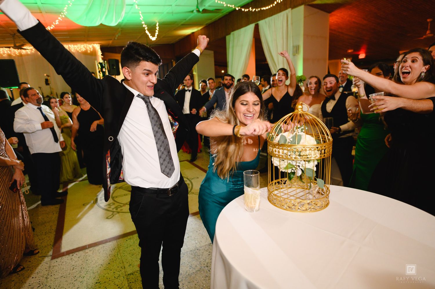 Couple celebrating at wedding reception with gold birdcage centerpiece and cheering guests in background.