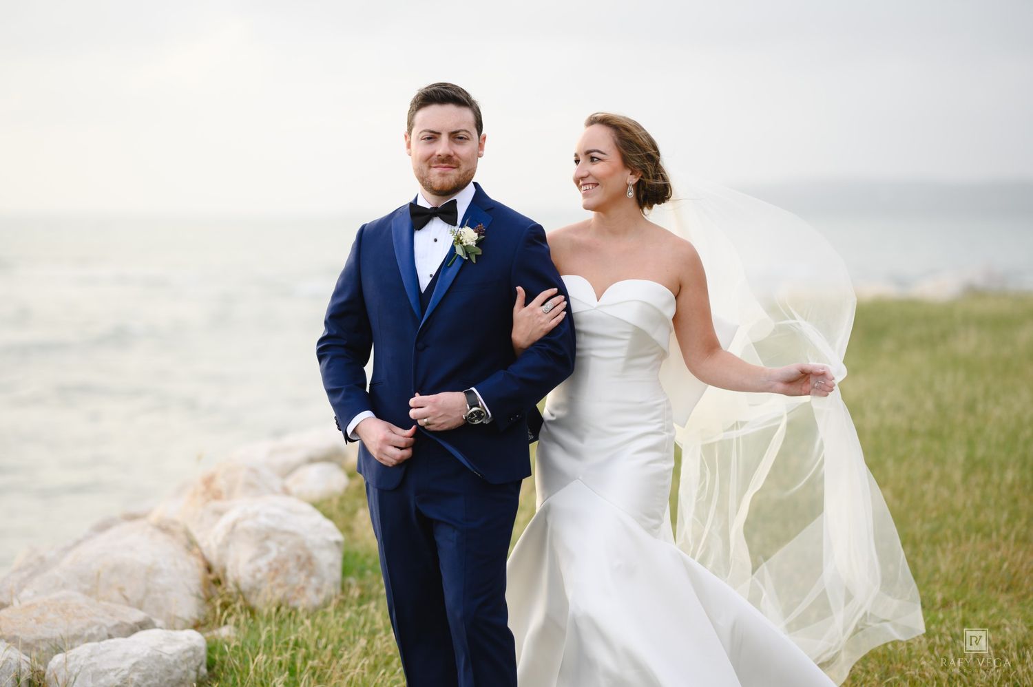 A bride and groom pose outdoors near rocky shoreline, her veil flowing in the wind, as he wears a navy suit.
