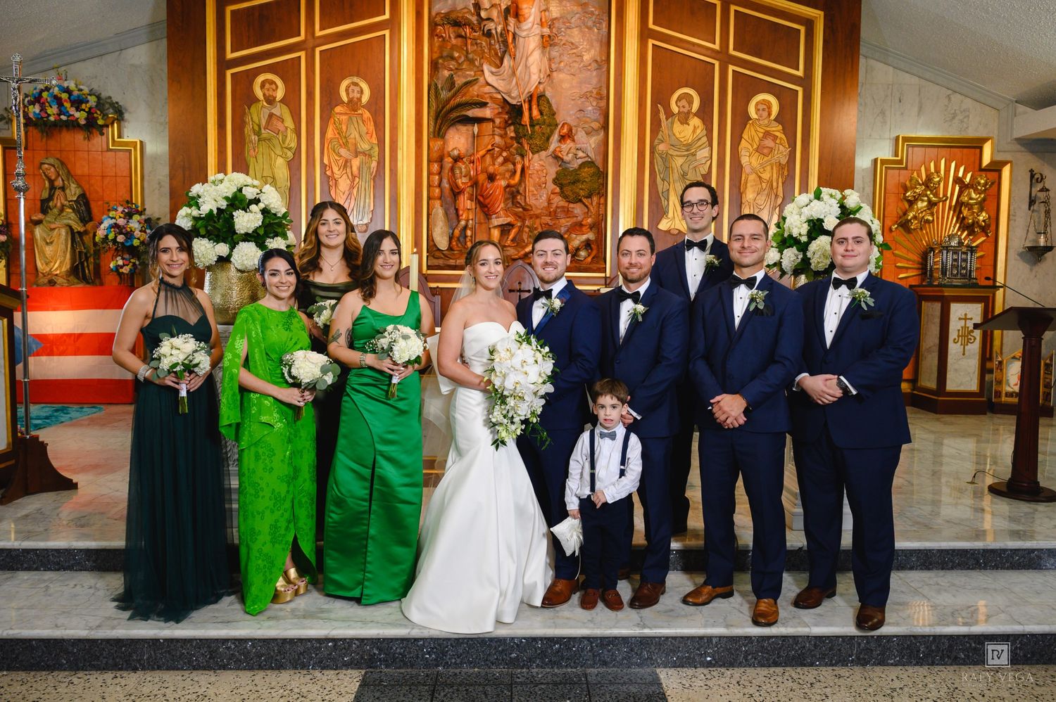 Wedding party poses in church with bridesmaids in green gowns and groomsmen in navy suits beside bride and groom.