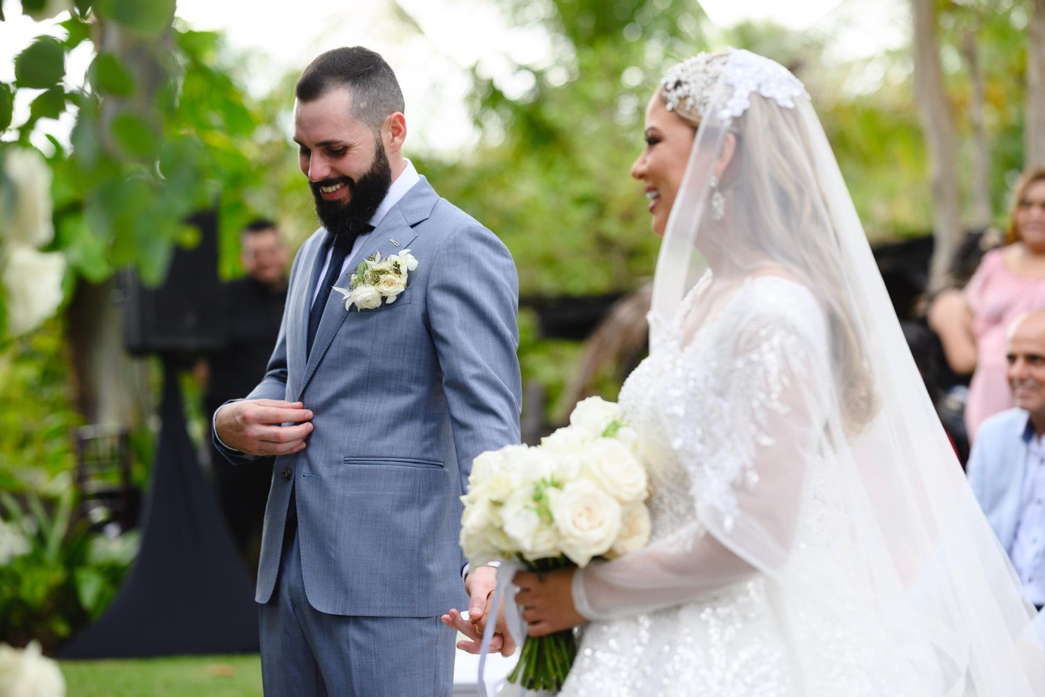 Bride in white lace gown and veil holds bouquet beside smiling bearded groom in gray suit at outdoor wedding.