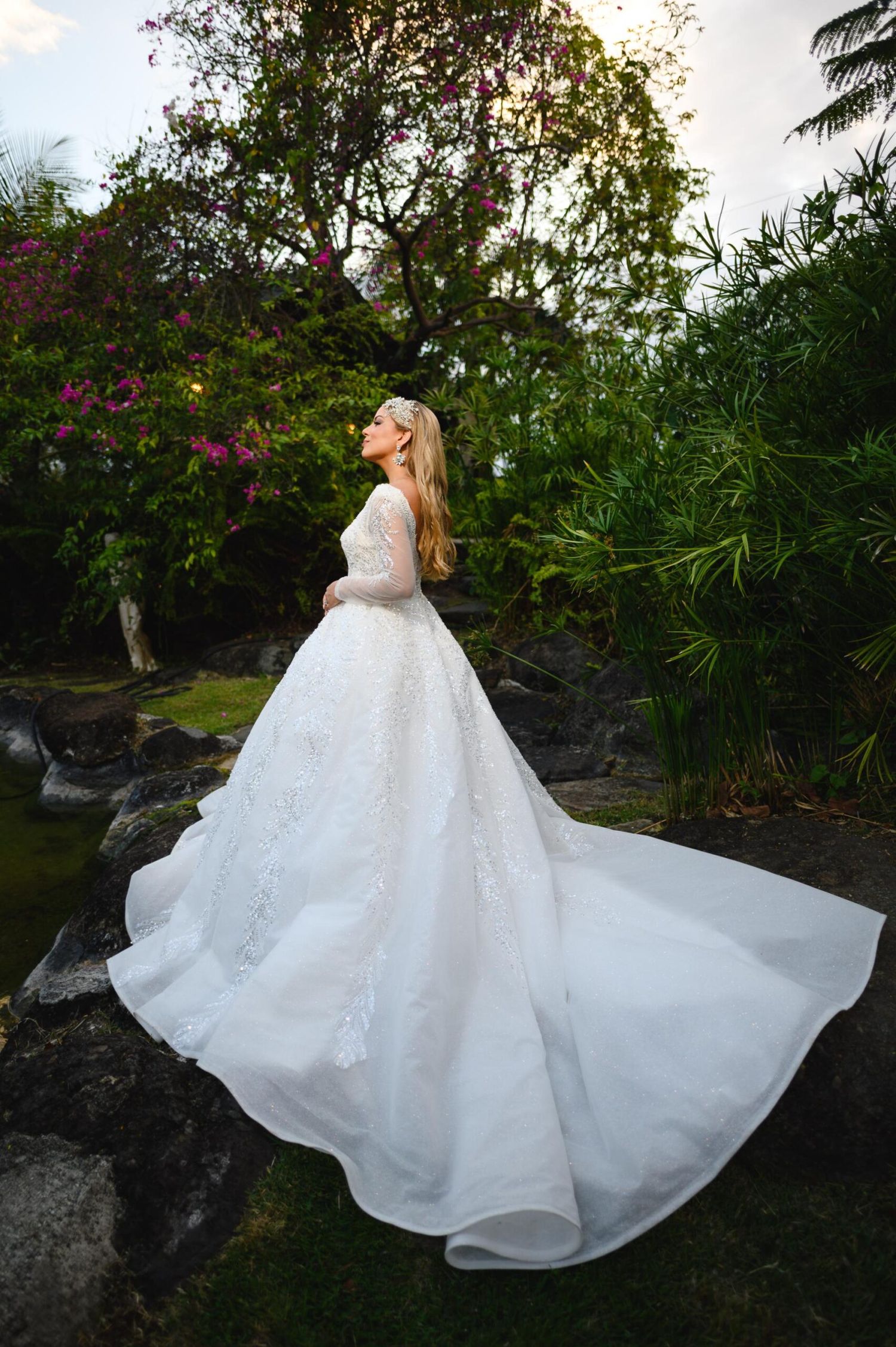 Bride in elegant lace ball gown with long train poses in lush tropical garden with pink flowers.