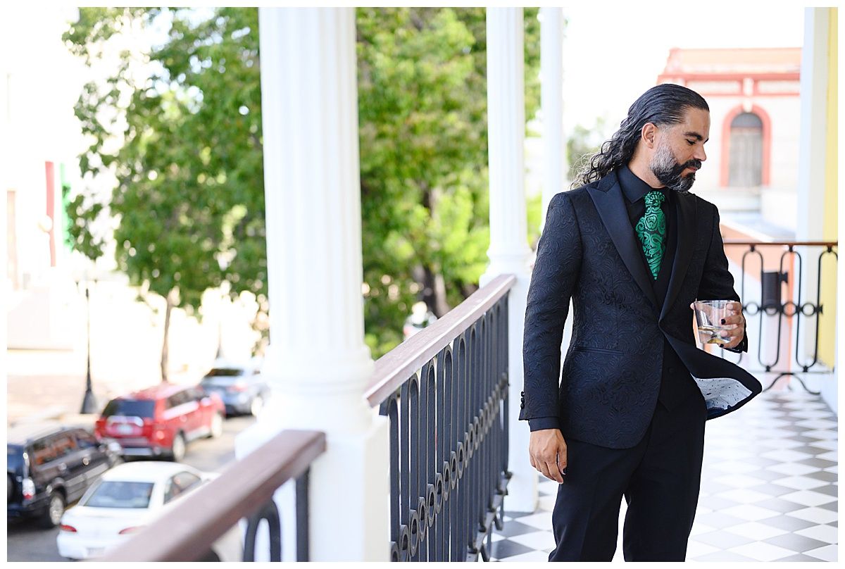 Stylish man in dark suit with green tie holding coffee cup on elegant white columned balcony.