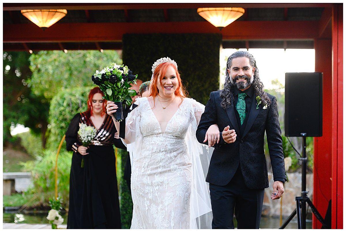 Radiant red-haired bride in lace gown walks with bearded groom after outdoor wedding ceremony recessional.