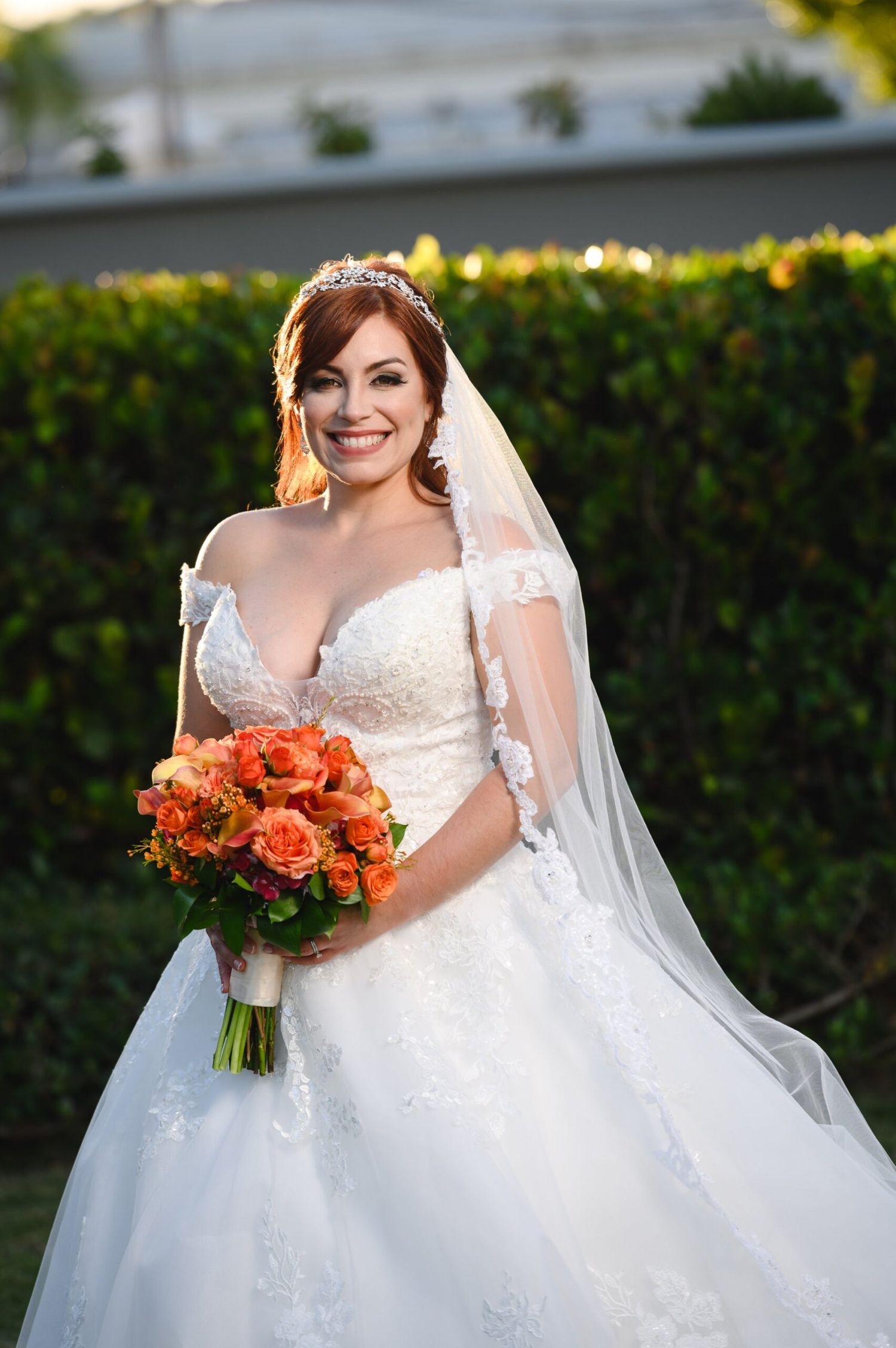 Smiling bride in white lace ball gown with veil and tiara holds orange rose bouquet outdoors.