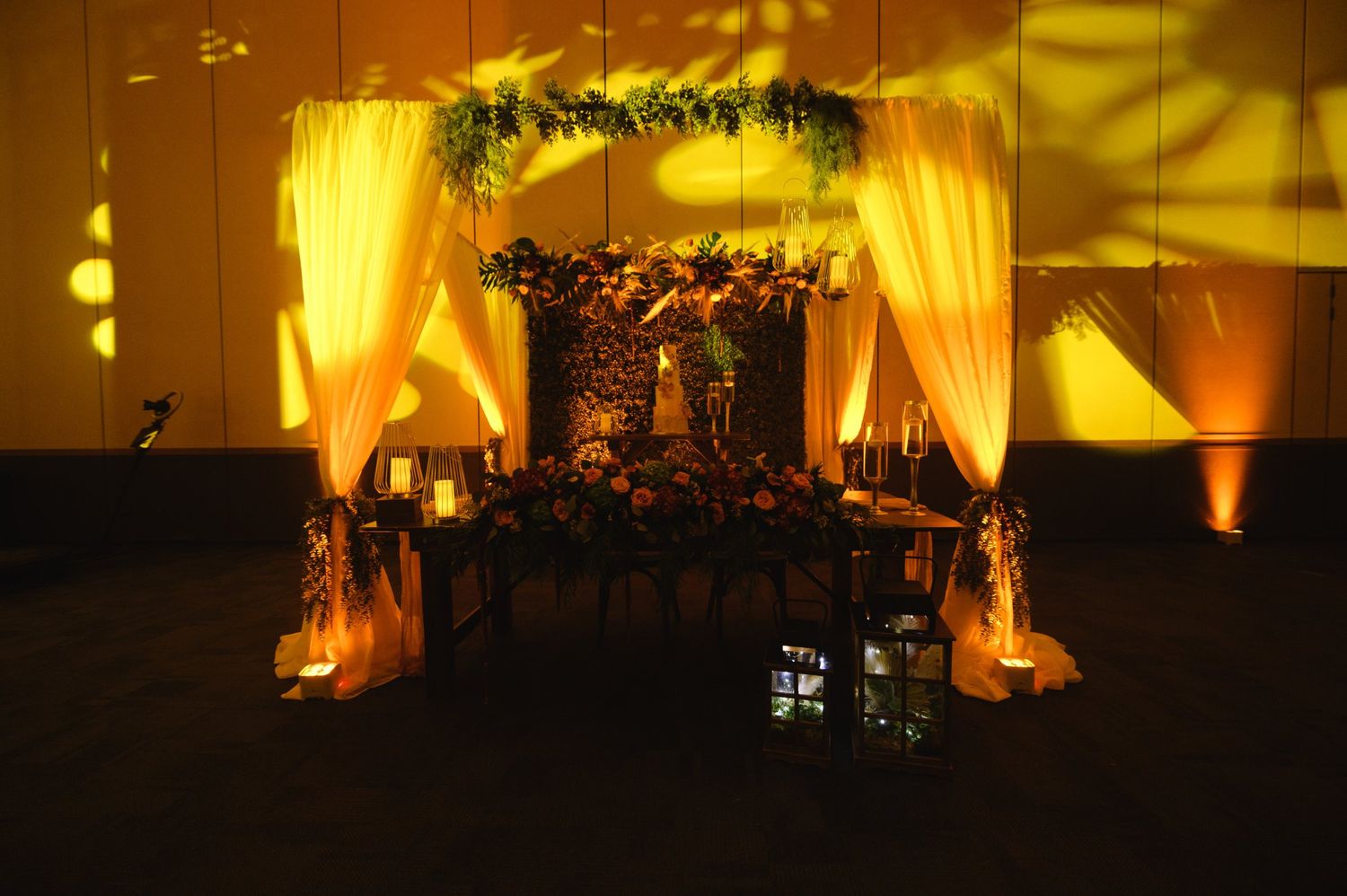 Elegant wedding sweetheart table with draped white curtains, floral arrangements, and warm golden uplighting in a dark reception hall.