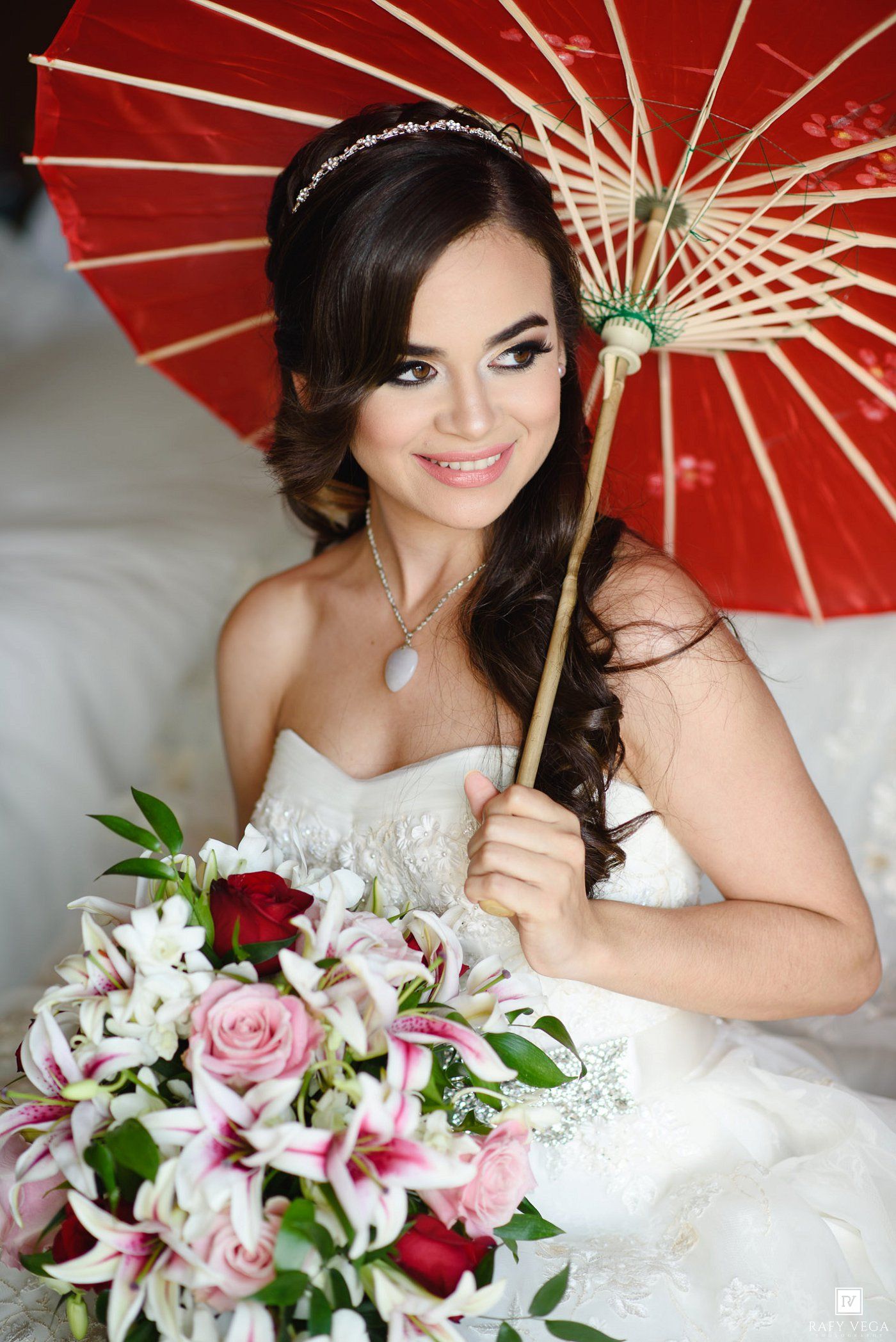 Smiling bride in white strapless gown holds red parasol and floral bouquet with pink roses and lilies.