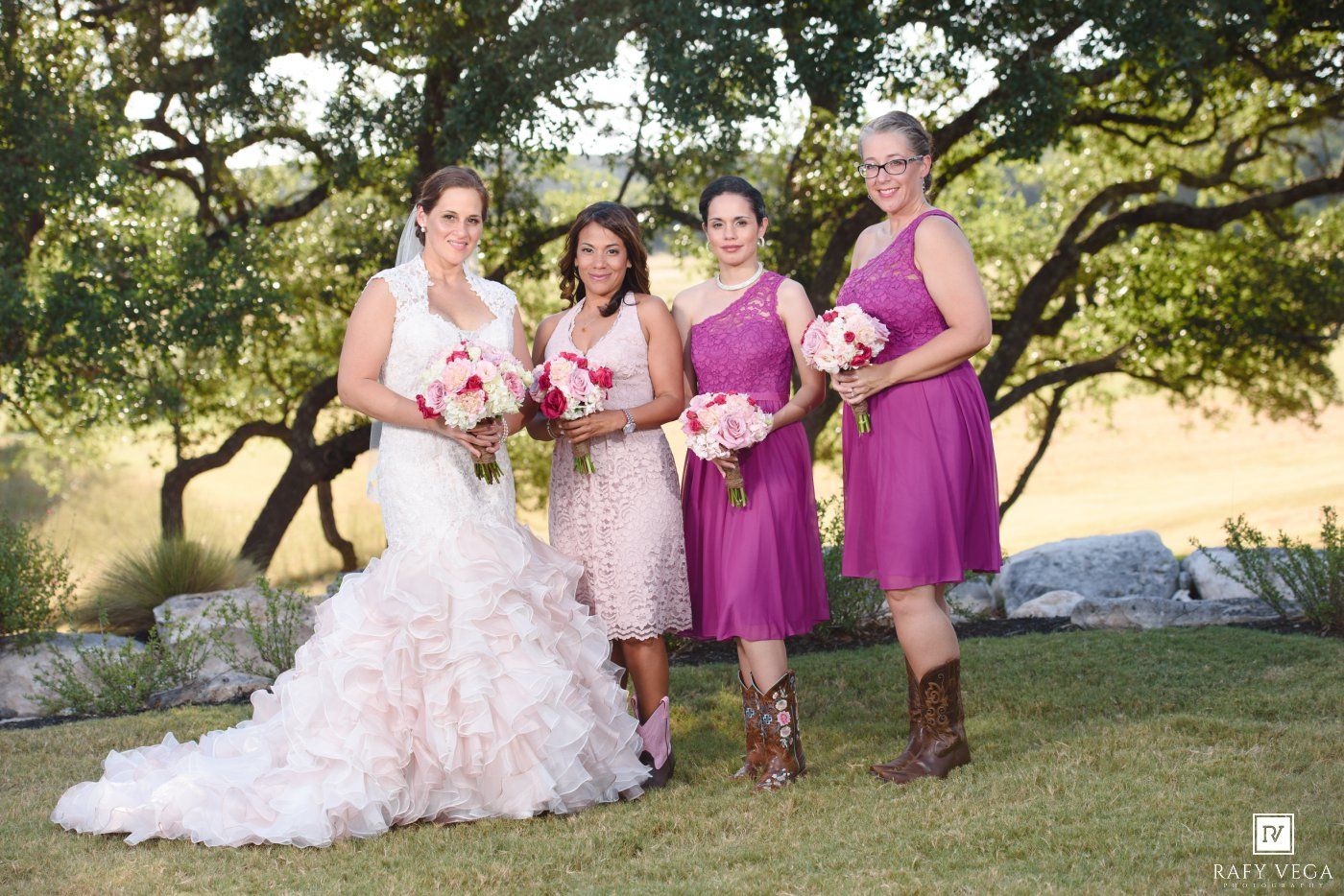 Bride in ruffled white gown poses with three bridesmaids in purple and pink dresses holding bouquets outdoors.