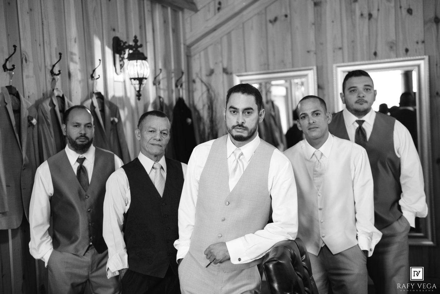 Black and white photo of a groom and groomsmen in vests posing together inside a rustic wooden venue before a wedding.