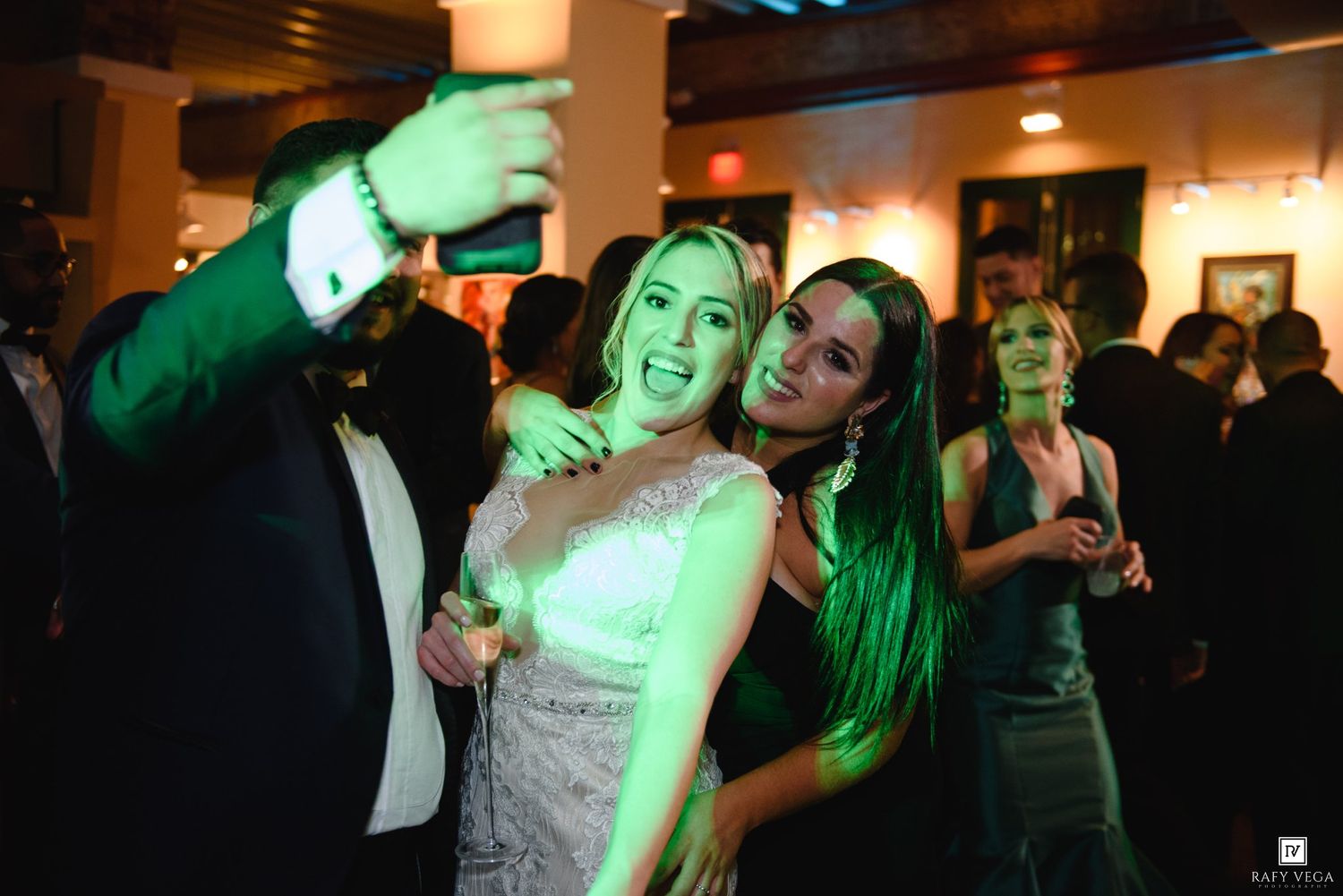 Two women smile and pose for a selfie at an elegant evening party with green lighting.