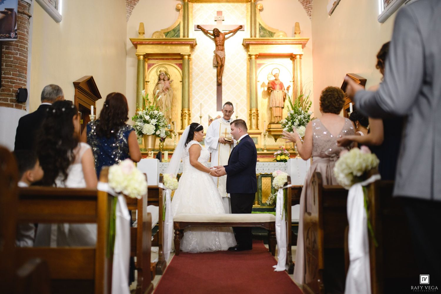 Bride and groom exchange vows at the altar of a decorated Catholic church surrounded by wedding guests.