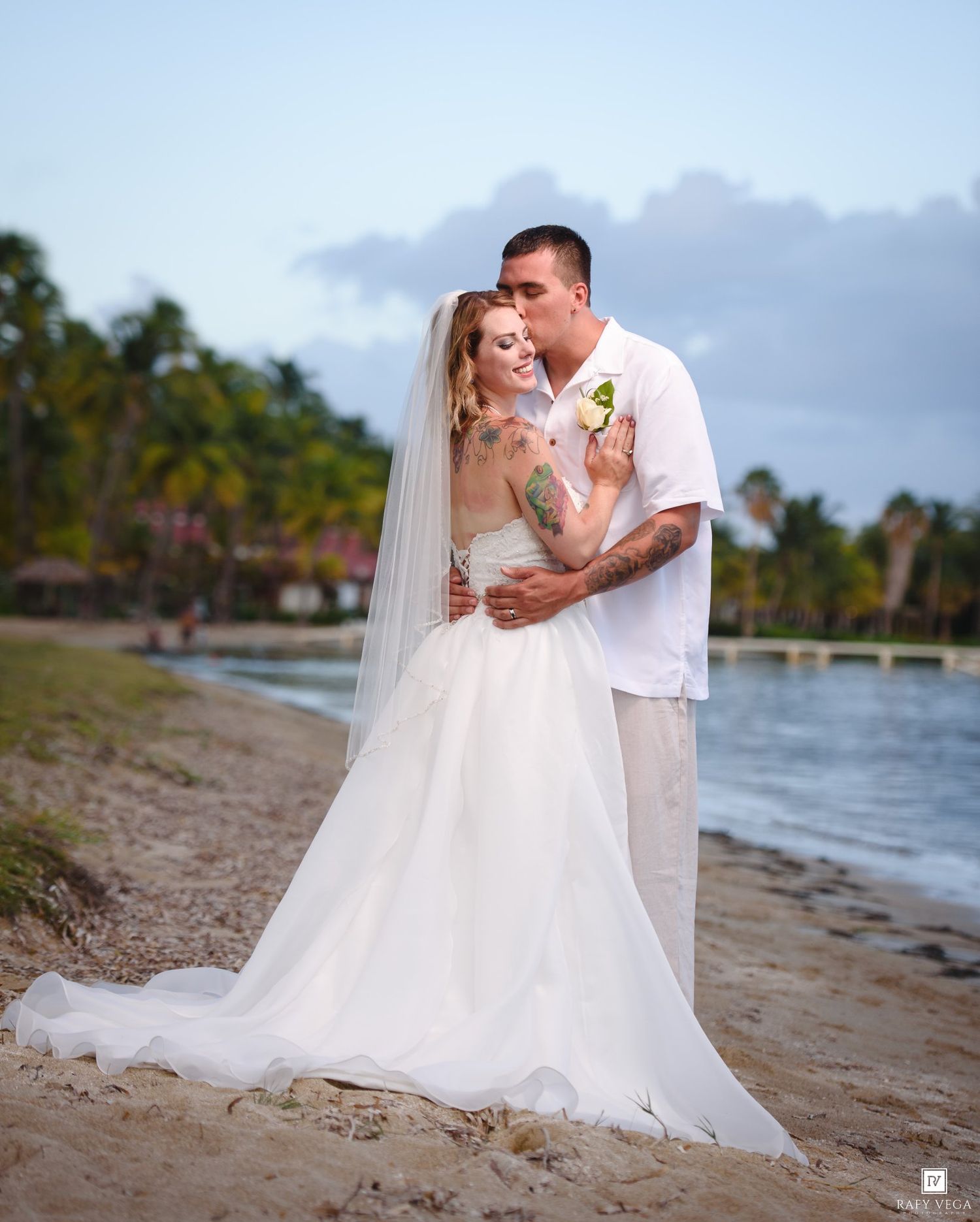 Bride and groom embrace on a tropical beach at sunset, bride in white gown with veil, groom in white shirt.