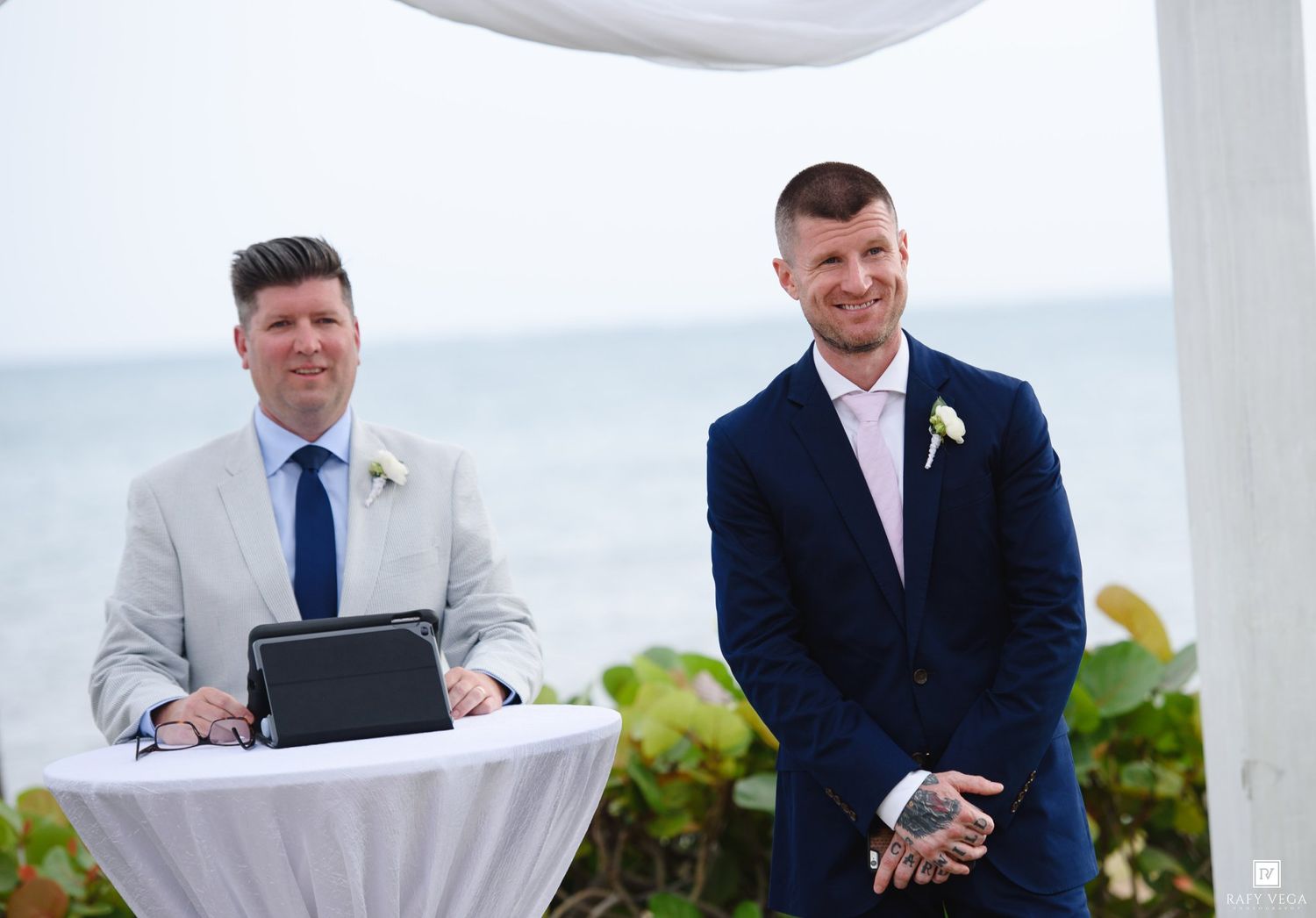 Two grooms stand at outdoor beach wedding ceremony, one in navy suit, one in light gray suit with blue tie.