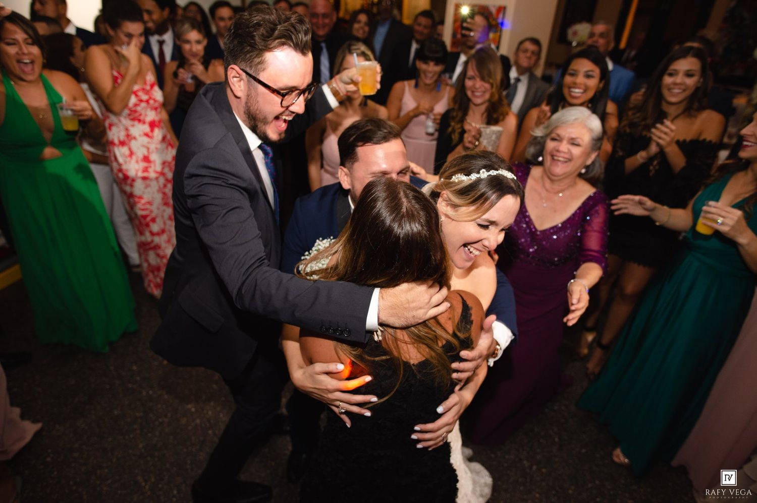 Joyful group hug on wedding dance floor surrounded by guests in formal attire at elegant reception.