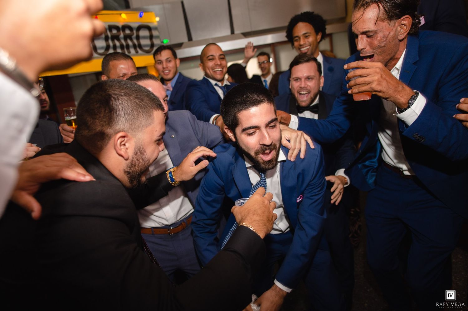 Groom in blue suit surrounded by cheering groomsmen celebrating at a lively wedding reception party.