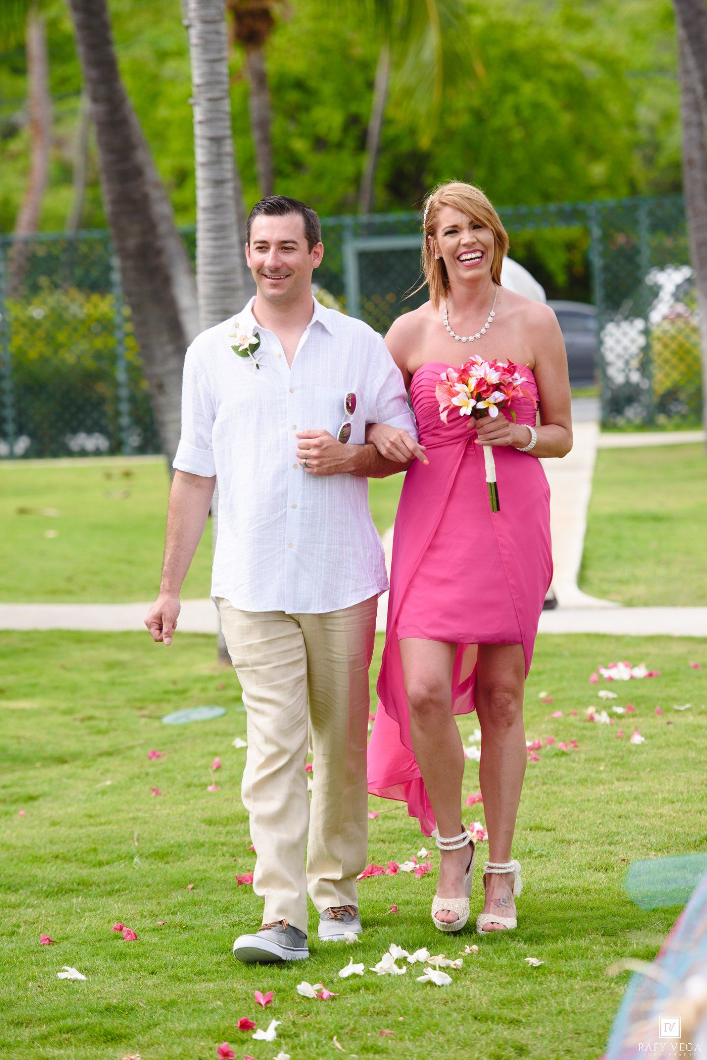 Smiling couple walks down outdoor aisle, woman in pink dress holding bouquet, man in white shirt, tropical wedding setting.
