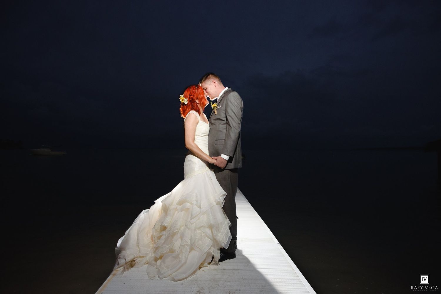 Bride and groom share a romantic kiss on a pier at night, with the ocean and dark sky behind them.