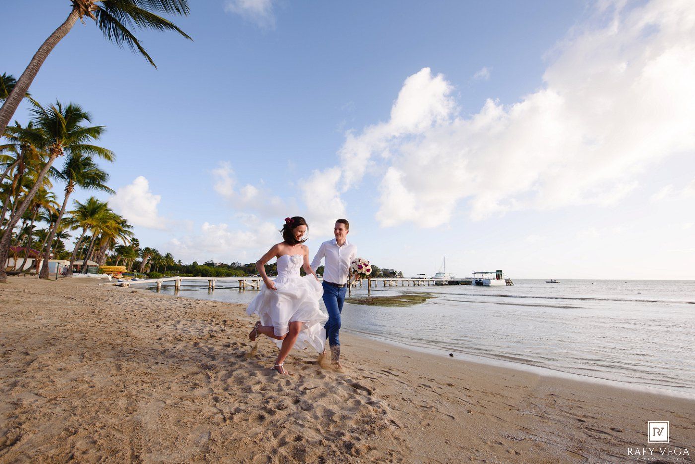 Bride and groom running joyfully on a tropical beach with palm trees and a pier in the background.