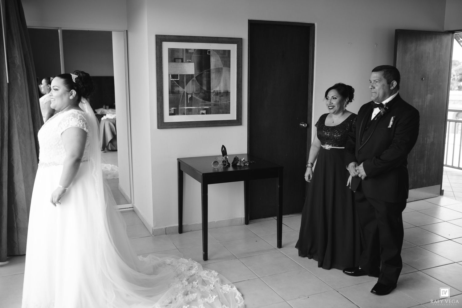 Black and white photo of a bride in a white gown with her parents watching proudly in a hotel room.