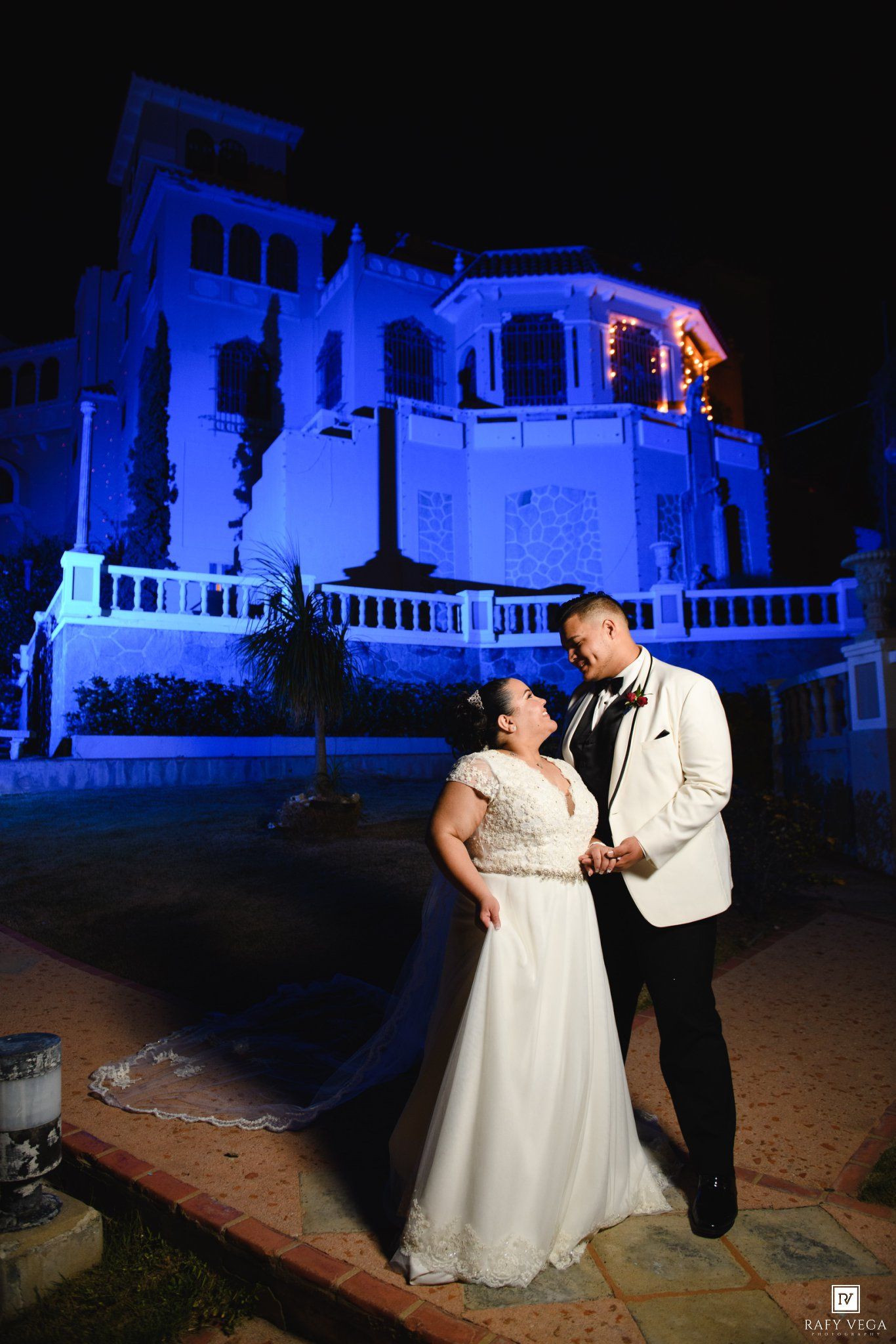 Bride and groom share a romantic moment at night outside a blue-lit historic building during their wedding celebration.