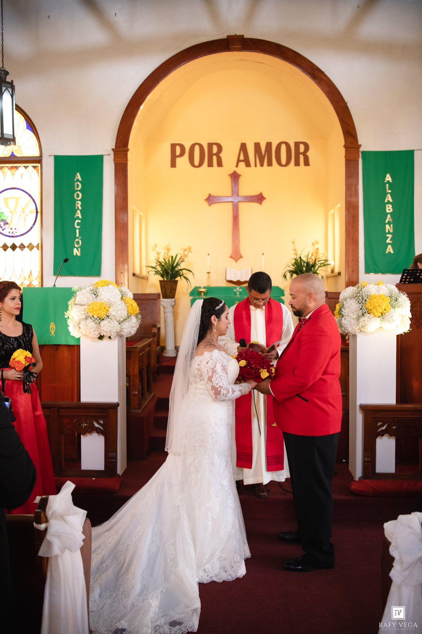 Bride and groom exchange vows at church altar with Por Amor sign, green banners, and floral arrangements.