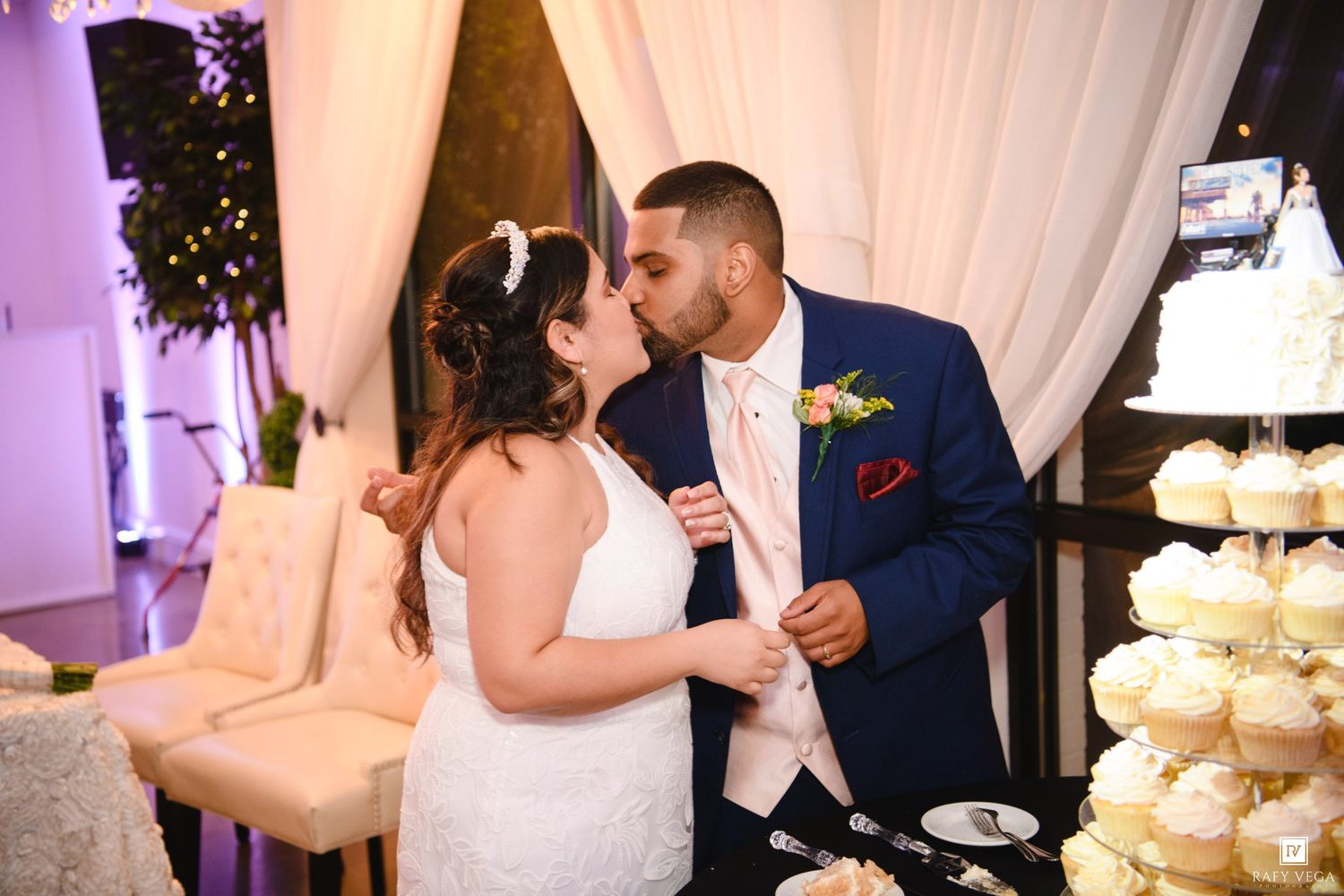Bride and groom share a kiss while cutting their wedding cake at an elegant reception with white drapes and purple lighting.